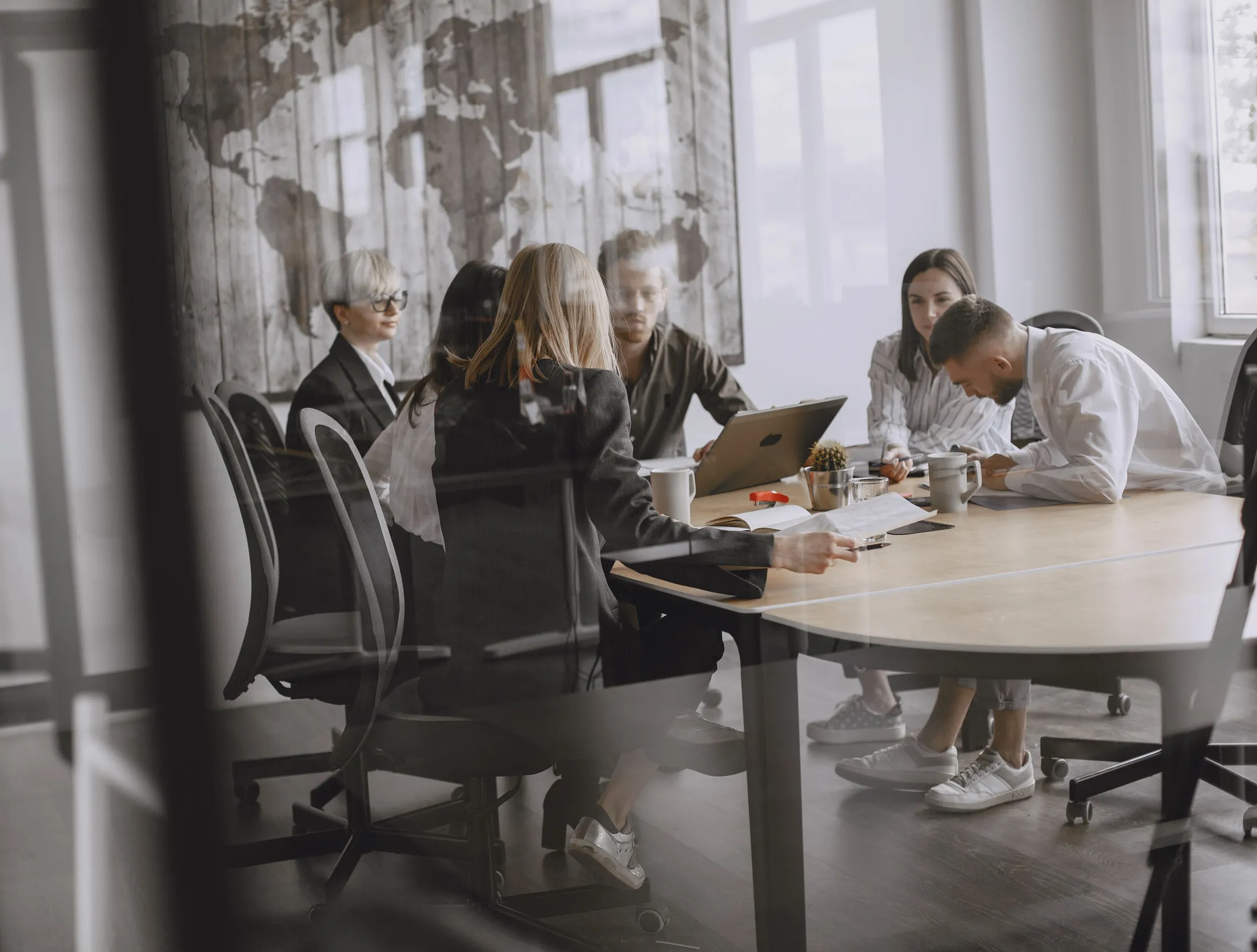 Profissionais reunidos em torno de uma mesa de trabalho, vistos através da divisória de vidro de um escritório contemporâneo.