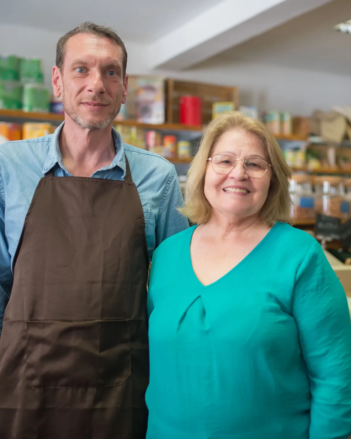 Dois comerciantes em uma mercearia de bairro, entre prateleiras de produtos e hortifrúti.