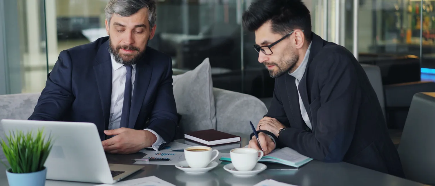 Dois profissionais analisando relatórios impressos e um notebook sobre a mesa.
